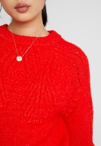 Pull en tricot rouge avec des motifs en relief et un col rond, complété par un collier en argent avec un pendentif rond.