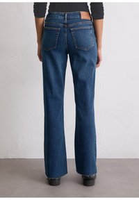 Sinine denim lõhekihtidega teksapüksid kõrge vöökohaga, viie tasku ja pruuni nahast seljataskuga. Kulunud hem ja sile tekstuur.