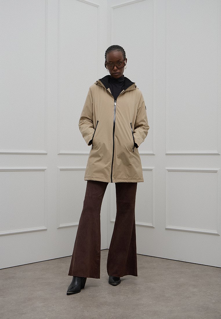 Beige waterdichte jas met een capuchon, voorrits en twee zijzakken, gecombineerd met bruine flarebroek en zwarte enkellaarsjes.