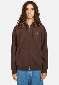 Bruine rits-hoodie van zachte stof, met voorzakken, een trekkoordkap en een klein logo op de borst. Gepaard met blauwe jeans.