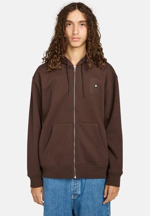 Braune Zip-Hoodie aus weichem Material, mit Fronttaschen, einer Kapuze mit Kordelzug und kleinem Logo auf der Brust. Getragen mit blauen Jeans.