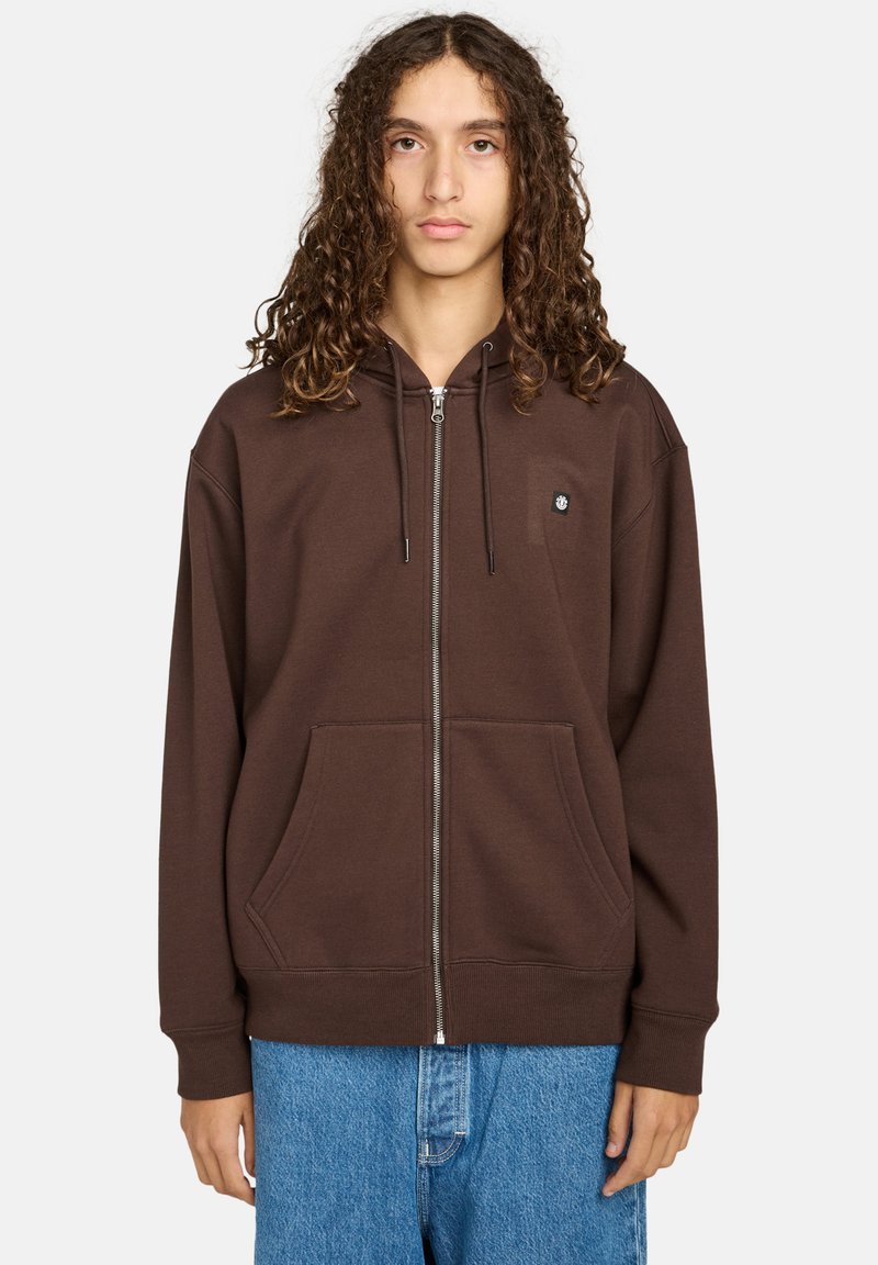 Braune Zip-Hoodie aus weichem Material, mit Fronttaschen, einer Kapuze mit Kordelzug und kleinem Logo auf der Brust. Getragen mit blauen Jeans.