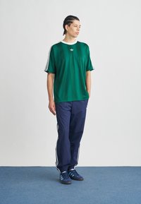 Mladić s pletenicama u zelenoj Adidas majici, tamnoplavim trenirkom i plavim tenisicama, stoji na plavom tepihu nasuprot bijele zidne površine.