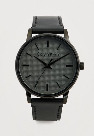 Montre analogique noire avec un cadran gris texturé, des index horaires noirs et un bracelet en cuir noir, dotée d'un boîtier en métal et d'un design minimaliste.