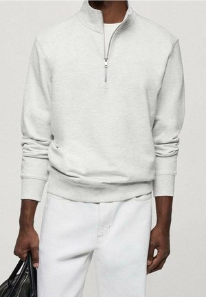 Mand iført en lysegrå kvart-lynlås trøje og hvide bukser, der holder en sort lædertaske i venstre hånd.