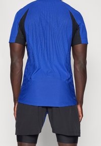 Blaues Sportshirt mit strukturiertem Design, kombiniert mit schwarzen Shorts. Kurze Ärmel und eine taillierte Silhouette betonen den Sportbekleidungsstil.