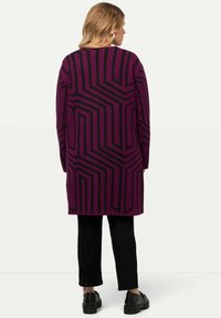 Maglione lungo e oversize viola con motivi geometrici neri. Presenta un colletto rotondo e spalle scese, abbinato a pantaloni e scarpe neri.