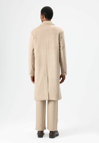 Beige wollen jas met een getailleerde pasvorm, voorzien van een verzonken kraag, lange mouwen en twee achterventilaties. Draagt over lichtbeige broek en bruine schoenen.