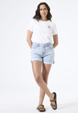 Vrouw met lang donker haar, gekleed in een wit t-shirt met een kleine afbeelding, lichtblauwe spijkershorts en bruine sandalen, staand met gekruiste benen.