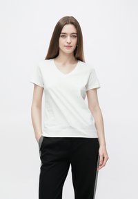 Weißes Baumwoll-T-Shirt mit V-Ausschnitt, kurzen Ärmeln und lockerem Sitz. Leicht strukturierte Oberfläche, kombiniert mit schwarzen Hosen mit Seitenstreifen.