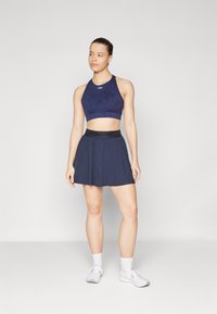 Tamno plavi sportski crop top uparen s odgovarajućom plisiranom suknjom, oboje izrađeno od lagane tkanine. Uključuje crni elastični pojas i bijele tenisice.