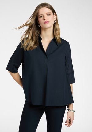 Kobieta o długich blond włosach, ubrana w luźną ciemnogranatową koszulę z rękawami 3/4 i dopasowane spodnie, pozująca na jednolitym białym tle.