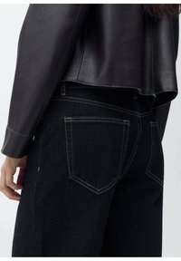 Sorte denim jeans med lige pasform, der har kontrasterende gule syninger og to lommer bagpå. Sammen med en mørk læderjakke.