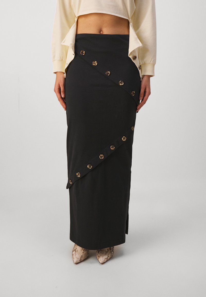 Jupe maxi noire avec des détails diagonaux à l'avant, ornée de grands boutons ronds, fabriquée en tissu lisse, avec une fente sur le côté.