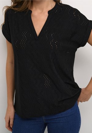 Vrouw met een zwarte blouse met oogjesstructuur, korte mouwen, een V-hals en omgeslagen manchetten, gecombineerd met een blauwe spijkerbroek.