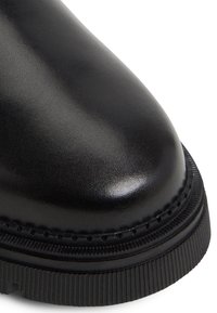 Musta nahkainen nilkkuri, jossa on pyöristetty varpaanosa, tekstuuripinta, koristeompelua reunassa ja uritettu kumipohja.