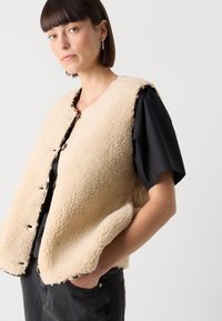 Beige Fleece-Weste mit rundem Ausschnitt, vorderen Knöpfen und seitlichen Taschen, über einem schwarzen kurzärmeligen Oberteil getragen. Strukturierter Stoff sichtbar.