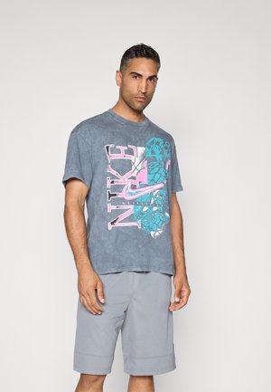 Camiseta de algodón gris con un gran gráfico de Nike en rosa y azul, cuello redondo y corte relajado. Combinada con pantalones cortos gris claro, de textura suave.