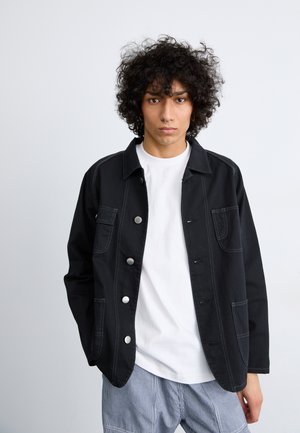 Black fabric jacket with a button-up front, two front pockets, and contrasting stitching, worn over a white t-shirt and striped pants.