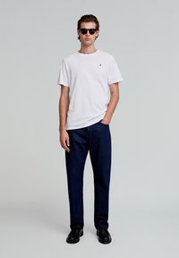 Witte t-shirt met ronde hals en klein zwart logo, gecombineerd met donkerblauwe rechtgesneden jeans en zwarte schoenen. Zonnenbril maakt de look af.