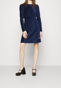 Abito lungo blu navy a maniche lunghe con texture a righe verticali, vita cinchata e orlo svasato, abbinato a scarpe nere con tacco a blocco.