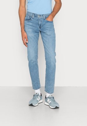 Mann trägt hellblaue Jeans, weiße Socken und hellblaue New Balance Sneakers, steht mit einer Hand in der Tasche vor weißem Hintergrund.
