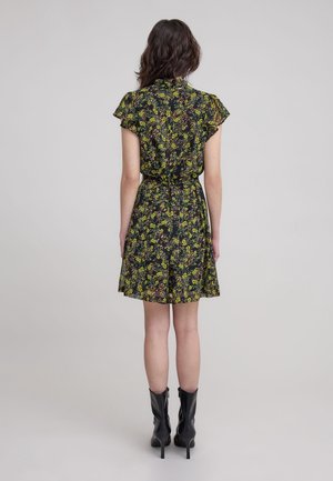 Schwarzes kurzes Kleid mit floralem Muster in Gelb und Grün. Hat kurze Ärmel, einen ausgestellten Saum und eine gebundene Taille. Trägt dazu schwarze Stiefeletten mit Absatz.