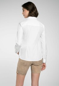 Witte langharige blouse met een getailleerd ontwerp, contrasterende stiksels en knoopaccenten op de manchetten. Draagt men met tan kleurige shorts van textuur stof.
