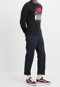 Felpa nera con grafica rossa "New Balance Classic", pantaloni blu navy e sneakers color marrone scuro. La felpa ha polsini e bordi a costine.