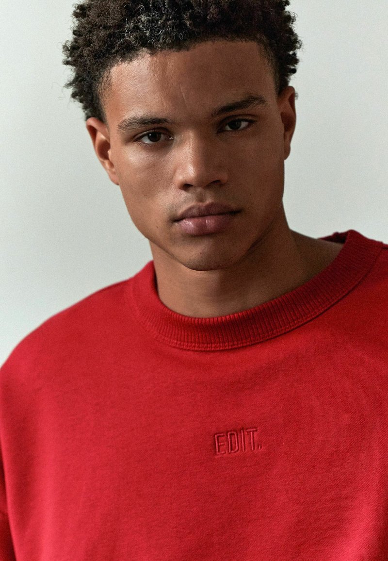 Rød sweatshirts med rund halsudskæring, prydet med broderet tekst "EDIT." på brystet. Blødt stof og afslappet pasform.