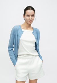 Cardigan bleu clair léger avec des boutons, porté sur un débardeur blanc et un short crème. Accent mis sur les textures douces et un design décontracté.