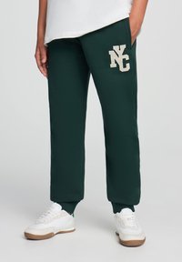 Zielone dresy o luźnym kroju, z beżowym logo "NY" po lewej stronie i ribbowanymi ściągaczami, w zestawie z białymi trampkami.