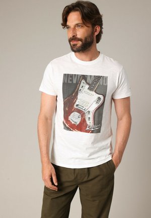 T-shirt en coton blanc avec un motif de guitare en rouge et gris, portant le texte "NEVER" et le nom de la marque "Deluxe" en bas. Manches courtes.
