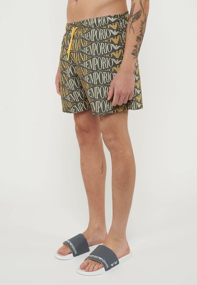 Uomo che indossa pantaloncini da bagno Emporio Armani neri e gialli con motivo a fantasia e ciabatte Emporio Armani grigie, in piedi davanti a uno sfondo bianco.