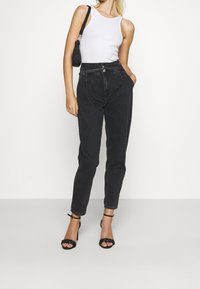 Zwarte high-waisted jeans met een taps toelopend been, voorzien van voorzakken en een ritsluiting. Gepaard met een witte mouwloze top en zwarte hakschoenen.