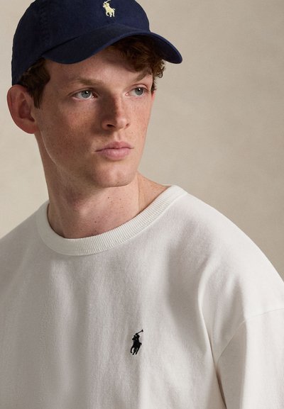 Νέος άνδρας με ελαφριές φακίδες που φοράει λευκό πουλόβερ με στρογγυλή λαιμόκοψη και σκούφο μπέιζμπολ σε σκούρο μπλε, και τα δύο με μικρό λογότυπο αλόγου και αναβάτη.