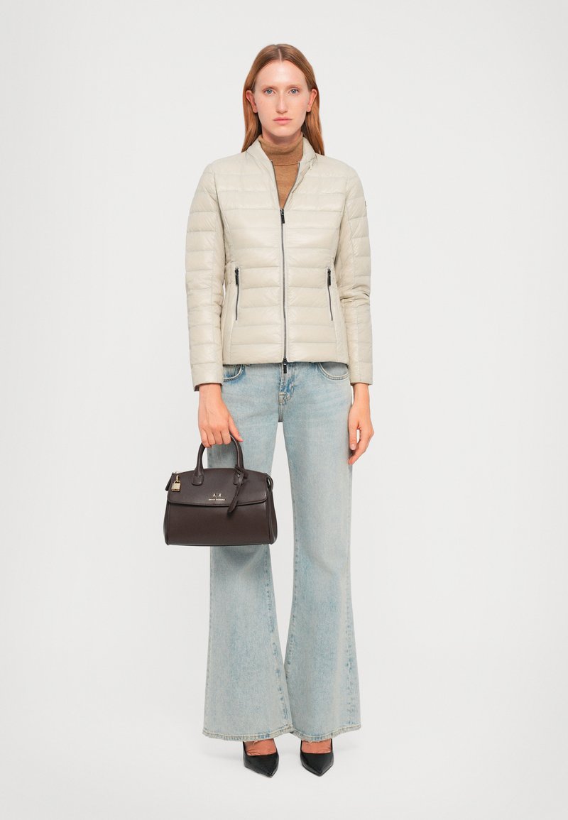 Veste matelassée beige avec un col montant, fermeture éclair et poches latérales. Associée à un jean flare bleu clair et un petit sac à main marron.