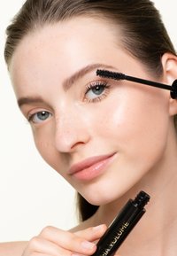 Un tubo di mascara nero tenuto da una persona con pelle liscia e lentiggini. Presenta un pennello spesso con setole nere per l'applicazione.