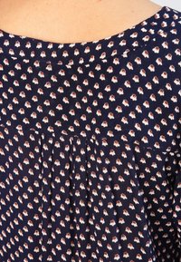 Donkerblauwe blouse met kleine witte en rode bloemprint, voorzien van een geplooid ontwerp aan de achterkant en een ronde halslijn. Zachte stofstructuur.