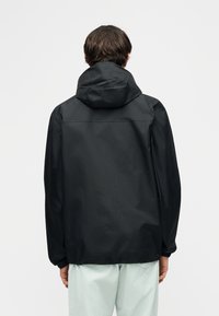 Άτομο φορώντας μαύρο αδιάβροχο με κουκούλα, φαίνεται από πίσω, συνδυασμένο με ανοιχτόχρωμο παντελόνι πάνω σε απλό φόντο.