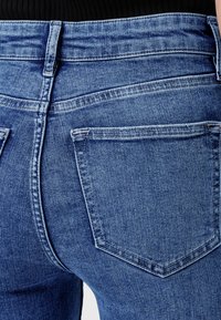 Spijkerbroek met een klassieke blauwe wassing, voorzien van een hoge tailleband, achterzakken en subtiele stikdetails. De textuur lijkt glad en duurzaam.
