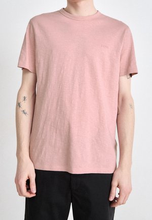 Osoba nosząca prostą jasnoróżową koszulkę z krótkim rękawem z małym wytłoczonym logo "BOSS", czarne spodnie i widoczne tatuaże na obu ramionach.