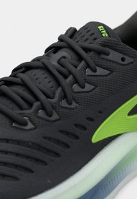 Juoda sportinė avalynė su tekstūruotu audinio viršumi, žaliais akcentais, tiesiomis raištelėmis ir permatomu padu. Šonuose yra ventiliacijos angos.