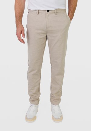Mann trägt beige Slim-Fit-Hose, weiße Sneaker und ein weißes T-Shirt, mit einer Hand in der Tasche und der anderen locker seitlich.