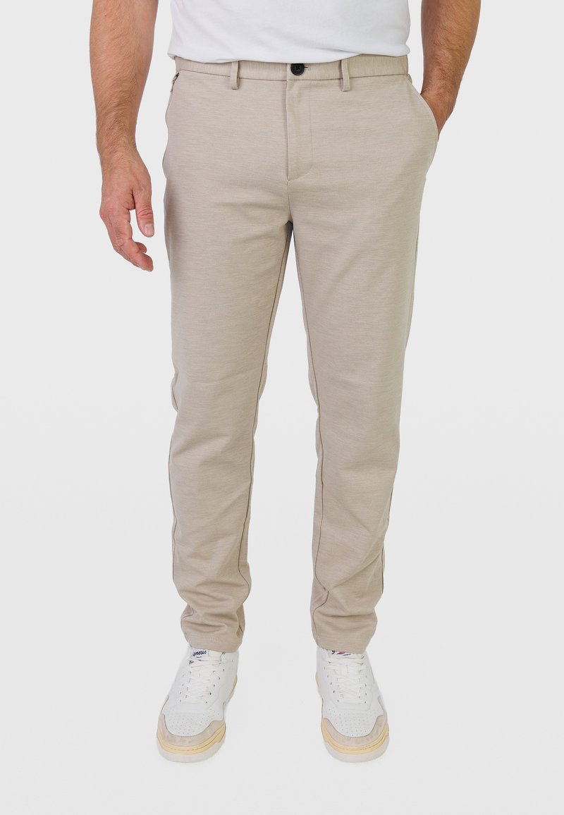 Homme portant un pantalon beige ajusté, des baskets blanches et un t-shirt blanc, avec une main dans la poche et l'autre détendue le long du corps.