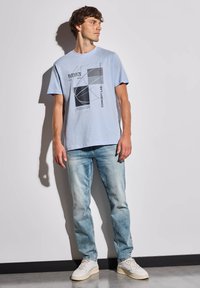 T-shirt en coton bleu clair avec impression graphique en noir et gris, associé à un jean délavé et des baskets blanches, offrant une coupe décontractée.