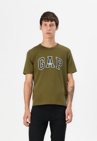 Olivgrünes Baumwoll-T-Shirt mit einem auffälligen navy-blauen "GAP"-Logo mit weißen Akzenten. Kurze Ärmel und Rundhalsausschnitt. Getragen mit schwarzen Hosen.