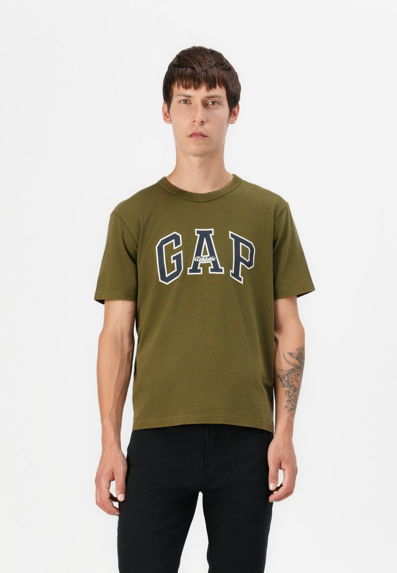 Olivgrünes Baumwoll-T-Shirt mit einem auffälligen navy-blauen "GAP"-Logo mit weißen Akzenten. Kurze Ärmel und Rundhalsausschnitt. Getragen mit schwarzen Hosen.
