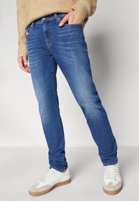 Blå denimjeans med slim fit, som har en subtil blekning, fem fickor och knappgylf, ihop med vita sneakers och en beige tröja.
