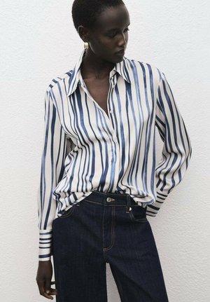 Camicia con bottoni a righe blu e bianche, con colletto, maniche lunghe e vestibilità ampia. Abbinata a jeans di denim scuro.